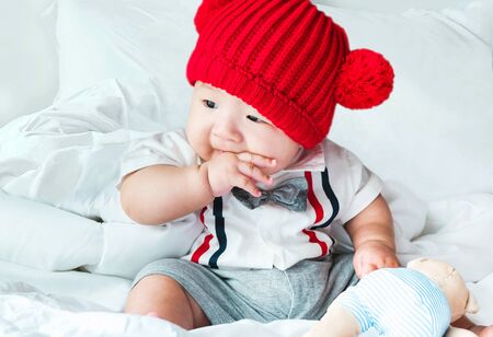 Portrait of a newborn Asian baby boy, Charming child 5 month old wore a suit and a red wool hat sitting in bedroom sucking his finger,fat baby cute and smilingly with a doll,soft selective focusの写真素材