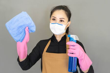 Asian Woman housewife in brown apron wore face mask ,hold the cleaning spray bottle and the cloth is wiping the glass isolated on gray background,housework and household concept.の写真素材