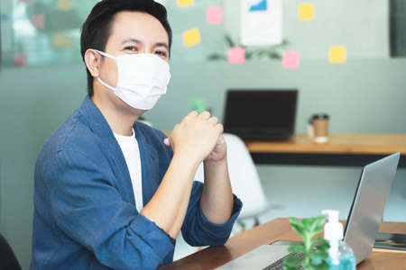 Young smart asian business man wearing medical face mask in modern office with laptop working under quarantine from coronavirus or COVID-19の写真素材