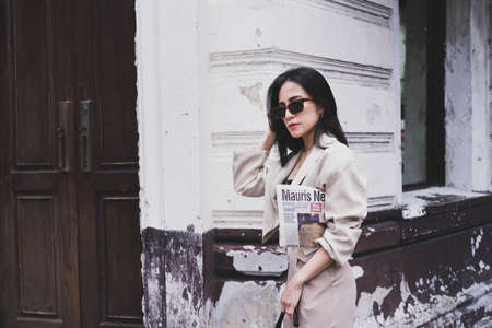 Asian pretty young hipster woman in trendy sunglasses in a vintage jacket in brown long pants with a...