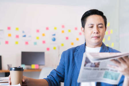 Young businessman sitting in office, reading newspaper, having coffeeの写真素材