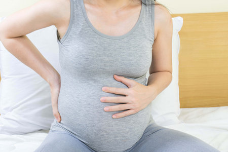 Close up of pregnant woman with back pain and stomachache sitting on bed at homeの写真素材