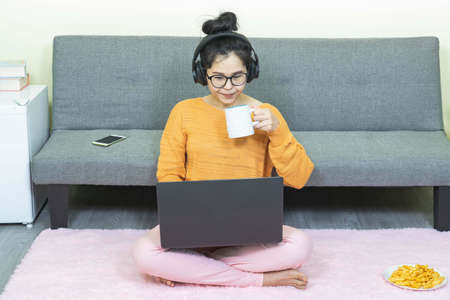 Smiling young asian woman enjoying morning coffee using laptop sitting at home. Home office concept.