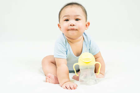 childhood and Childcare concept Portrait of cute little 5 months old  asian newborn baby boy wearing blue bodysuit sitting on white beedsheets with sippy cup.Selective focus.の写真素材