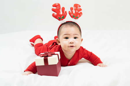 New year and Christmas concept with 5 months old cute asian newborn baby boy,smilimg, wearing christmas antlers of a deer,red clothes lying on the blanket with gift box.Gifts for kids. Holiday season.の写真素材