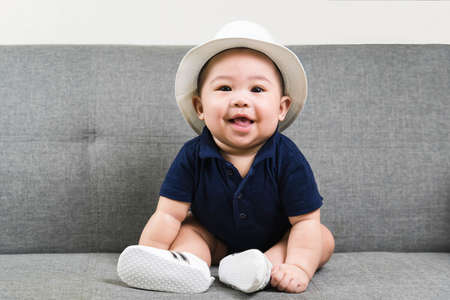 Adorable baby boy  first sitting on sofa. Cute infant Asian about 5-6 months old wearing blue t-shirt,white hat and shoes smiling and looking at camera.の写真素材