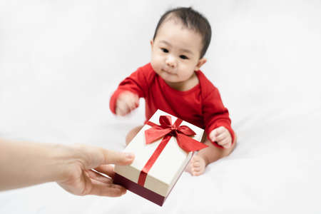 Cute asian baby in red bodysuit get gift box sitting on white blanket at home.Give Year present to child,kid,toddler.Concept holiday Christmas, happy new year, infants, childhood.selective focus.の写真素材