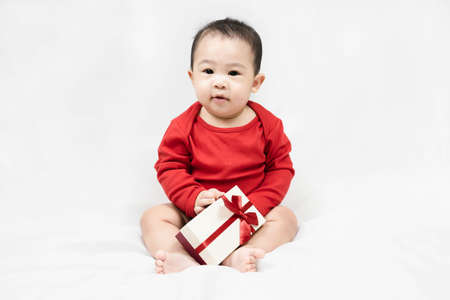 Happy infant asian baby boy toddler in red bodysuit holding gift box sitting on white background.Concept holiday Christmas, happy new year, infants, childhood.の写真素材