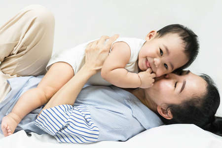 Selective focus Beautiful mom kissing on the cheek newborn with love and caring, Mother showing love to toddler with kiss on cheek.infant baby lying on bed with asian mom.の写真素材