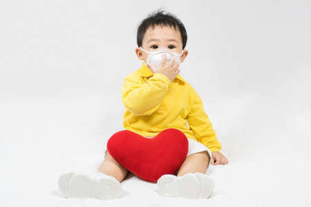 Asian cute baby boy about one year old in a protective mask and yellow shirt with red heart shape pillow, coronavirus and covid-19 pandemic concept.Selective focus.の写真素材
