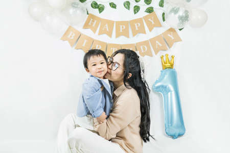 Asian baby boy celebrating first birthday,A mother hug and kiss her cheek son smiling on a floor with a minimal background,white balloons and birthday flag.Selective focusの写真素材