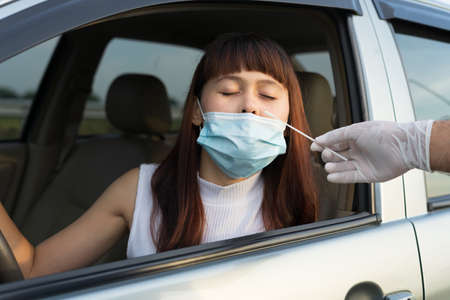 Woman sitting in car for medical worker through car window to perform drive-thru COVID19 test. Doctor or healthcare hand white glove with cotton swab making coronavirus test for young lady in her car.の写真素材