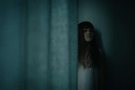 Female ghost in white dress in room. A young scary girl in an old white dress staring in to the camera ferociously with dark background. Asian woman make up ghost face at house. Scary horror concept.の写真素材