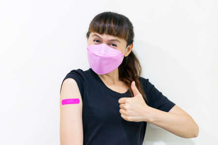 Asian woman wear face mask using adhesive bandage at with pink arm and showing thumb up gesture after injection vaccine. Covid 19 vaccine disease preparing.の写真素材