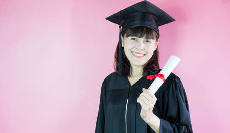 Happy graduate smiling she is in graduation cap and diploma in hand on pink background.