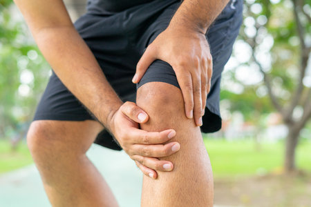 A young male in sports outfits black injured his knee during exercise in the park. Low section of sports man suffering from joint pain while standing on track during. Accident from exercise concept.の写真素材