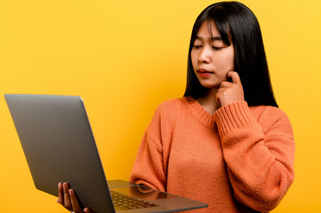 online communication concept asian girl use laptop at home yellow studio photo She is happy to spend her free time online while chatting with friends on social networks, shopping or working online.の写真素材