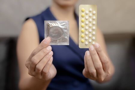 Woman hand holding contraceptive pills and condom sitting on sofa background,protection, safe sex,contraceptive means.の写真素材
