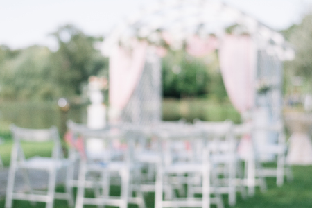Beautiful summer wedding ceremony outdoors. Decorated chairs stand on the grass. Wedding arch made of light cloth and white and pink flowers on a green natural background. White wooden doors, rustic style. Empty seats with blurred focusの写真素材
