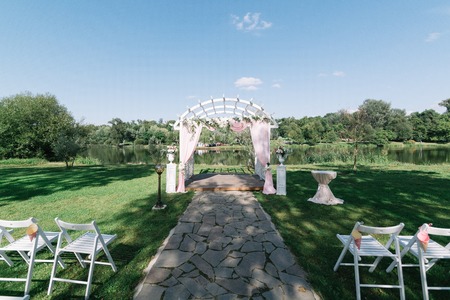 Beautiful summer wedding ceremony outdoors. Decorated chairs stand on the grass. Wedding arch made of light cloth and white and pink flowers on a green natural background. White wooden doors, rustic style. Empty seats.の写真素材