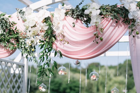 Beautiful decoration for summer wedding ceremony outdoors. Wedding arch made of light cloth and white and pink flowers on a green natural background. White wooden doors, rustic style.の写真素材