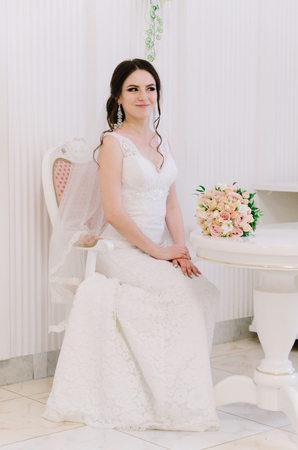 Beautiful bride perfect style. Wedding hairstyle make-up dress and bride's bouquet. Young attractive bride on chair in white room at studio.の写真素材