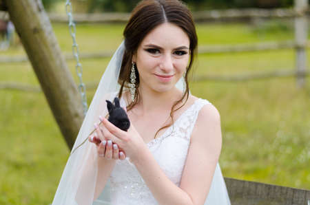 Young woman bride smiling and holding cute rabbit over park summer nature outdoor. White wedding dress, green background.の写真素材