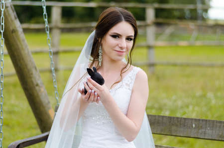 Young woman bride smiling and holding cute rabbit over park summer nature outdoor. White wedding dress, green background.の写真素材