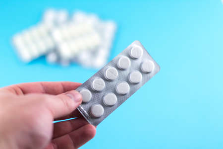 The man hand is holding a pack of white pills packed in blisters with copy space on blue background. Focus on foreground, soft bokeh.の写真素材