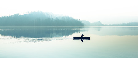 Fog over the lake. Silhouettes of trees. The man floats in a boat with a paddle. Filter softlight.の写真素材