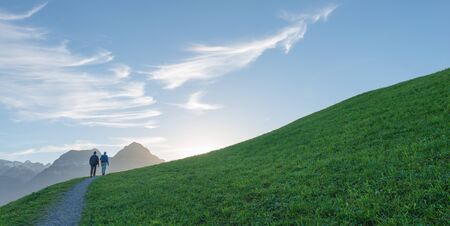 Couple goes up the mountain path. Alpine meadow with green grass. Blue sky with interesting clouds, sunset. Away view of tops of mountains in snow.の写真素材