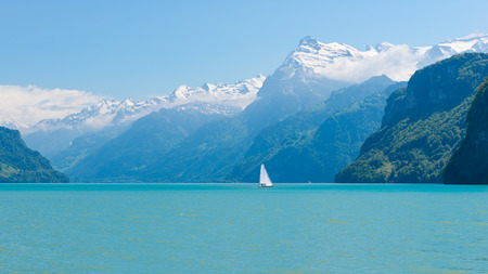 Magnificent panorama of mountains with snowy peaks. On  surface of lake sailboat sails. Cloudless blue sky and a few clouds in the mountainsの写真素材