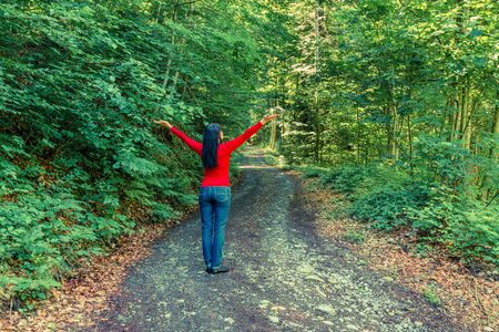 Woman in the forest on the edge. She raised her hands up in an appeal to nature. Sun rays in the trees. Further prospects of the road.の写真素材