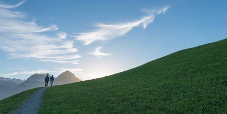Couple goes up the mountain path. Alpine meadow with green grass. Blue sky with interesting clouds, sunset. Away view of tops of mountains in snow.の写真素材