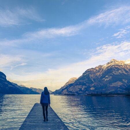 Woman goes on a wooden pier. Lake between the mountain ranges. Mountain landscape in the evening.の写真素材