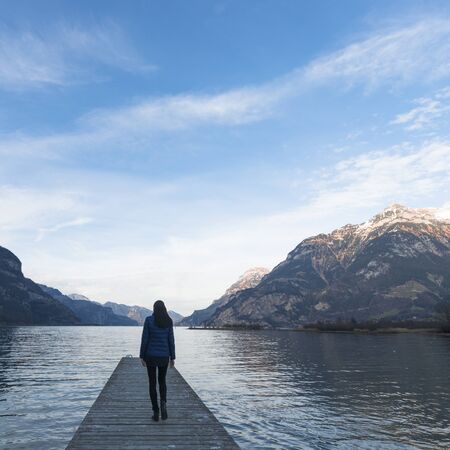 Woman goes on a wooden pier. Lake between the mountain ranges. Mountain landscape in the evening.の写真素材