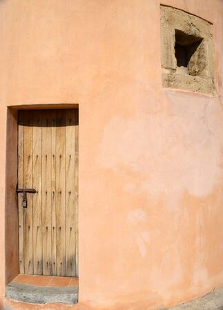 Rustic wooden door with iron nails and padlock. Small window over red earth wall.の写真素材