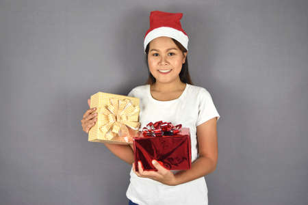 Portrait of happy woman wearing santa claus hat and holding gift box with ribbon on grey background....