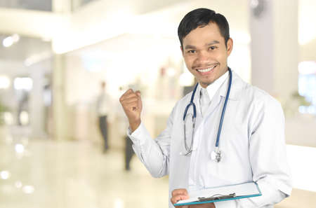 Young doctor smiling with stethoscope holding a clipboard and showing yes gesture of success on hospital blur background. copy space. healthcare and medicine concept.の写真素材