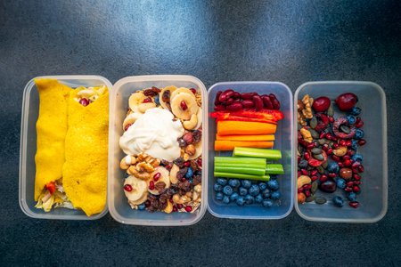 Boxes with healthy food full of colorful vegetables and fruits. Nuts, blueberries, yogurt, pomegranates, bananas, gluten-free pancakes, raisins, celery, carrots and peppers. Meals for the day.の写真素材