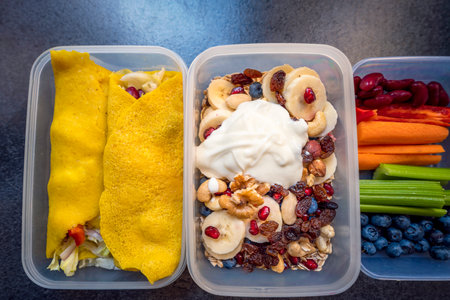 Boxes with healthy food full of colorful vegetables and fruits. Nuts, blueberries, yogurt, pomegranates, bananas, gluten-free pancakes, raisins, celery, carrots and peppers. Meals for the day.の写真素材