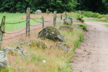 A rural gravel road. A fence made of wooden posts and barbed wire, and moss-covered boulders.の写真素材