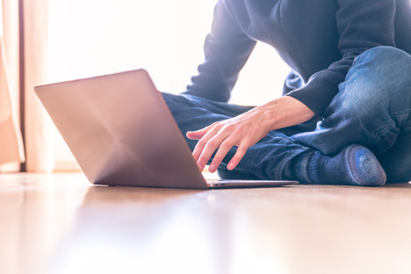 Young man with a laptop is working on the wooden floor, light comes in from the windowの写真素材