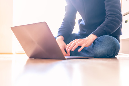 Young man with a laptop is working on the wooden floor, light comes in from the windowの写真素材
