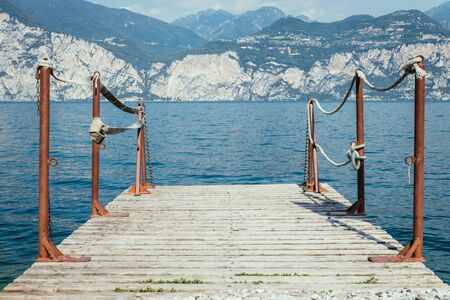 Wooden dock pier extending over blue lake water, mountains at lago di garda. Text space.の写真素材