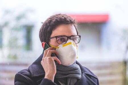 Young business man outdoors with a breathing mask and smartphone. Flue and corona season.の写真素材