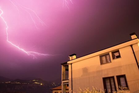 Lightning on the cloudy sky, mountains and lake, building in foreground, Italy