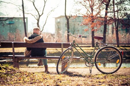 Back view of young woman with bicycle who is enjoying the sun on a park bench, spring timeの写真素材
