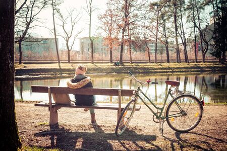 Back view of young woman with bicycle who is enjoying the sun on a park bench, spring timeの写真素材