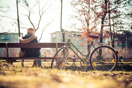 Back view of young woman with bicycle who is enjoying the sun on a park bench, spring timeの写真素材